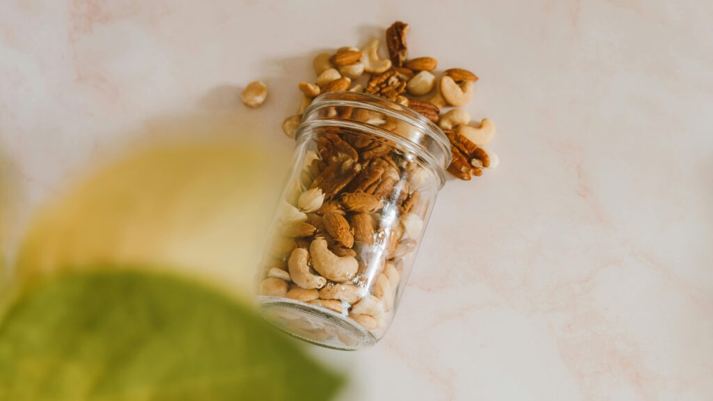 A glass jar spilling mixed nuts including cashews and almonds on a marble surface.